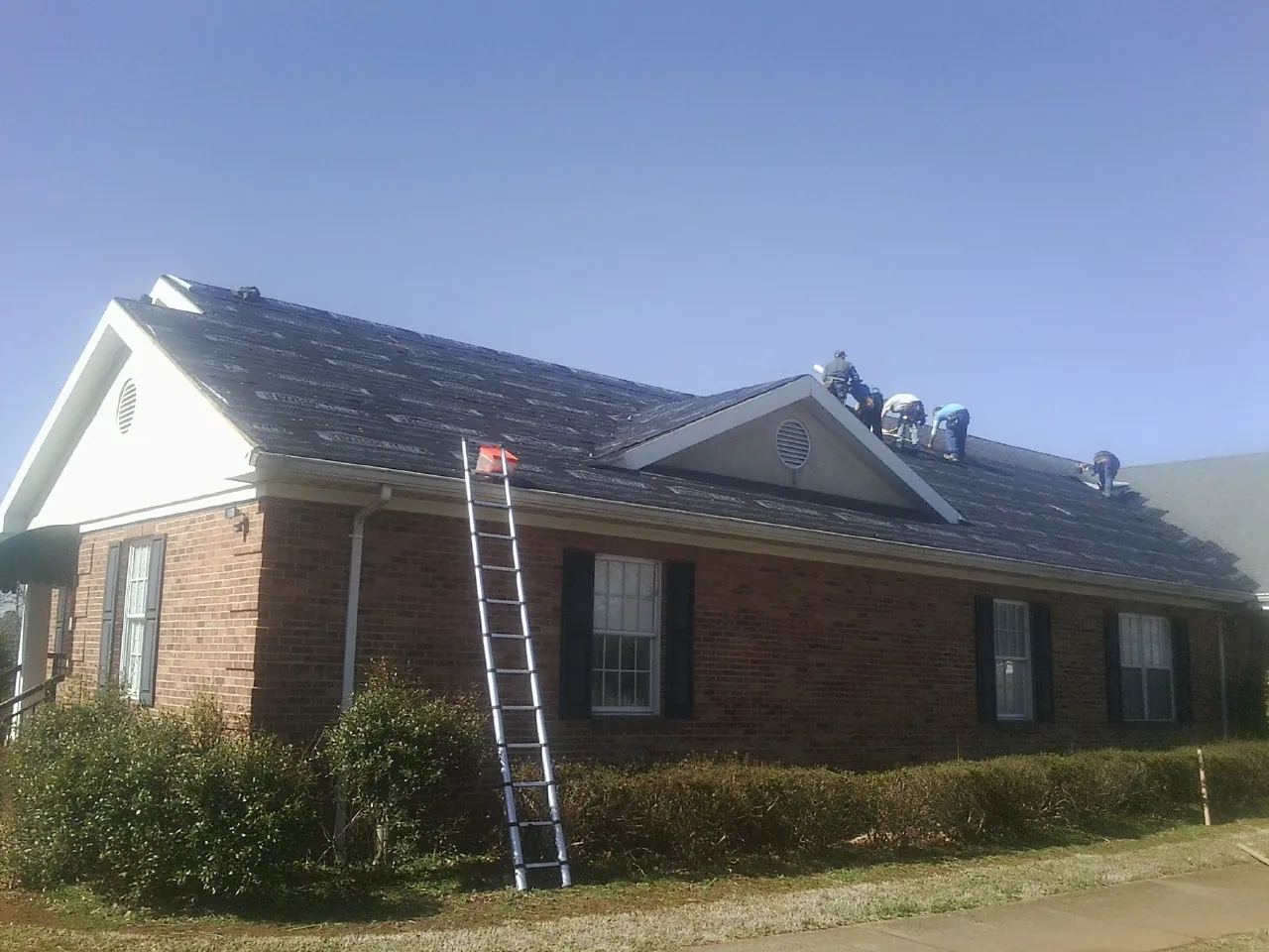 Crew tearing shingles off a brick ranch roof with a ladder set — Emergency Roof Repair work by our crew in Bluewater