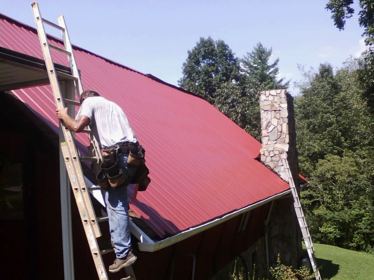 Roofer on a ladder beside a red metal roof and stone chimney — Soffit & Fascia Repair by Roofing Brothers in Bluewater
